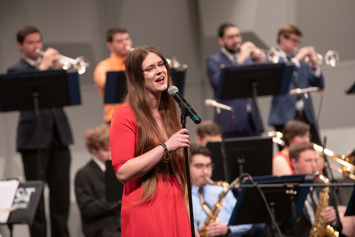 A woman in a red dress passionately sings into a microphone on stage, with a jazz band playing brass instruments in the background.
