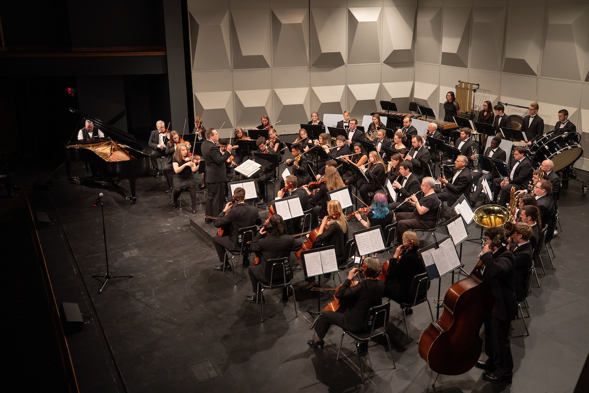 An orchestra performs on stage featuring violinists, a pianist, and percussionists.