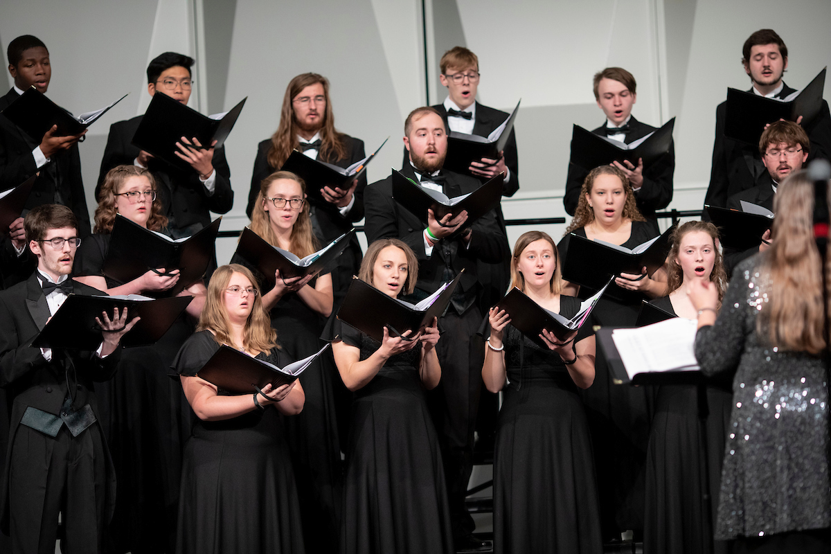 A choir of diverse singers dressed in formal black attire performs passionately, holding open music folders, led by a conductor in a sequined dress